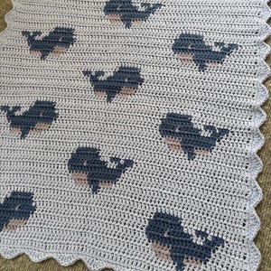 以下が含まれることがあります： 青とベージュの色合いのクジラの繰り返し模様が特徴の、水色の手編みブランケット。ブランケットはスカラップエッジで、手作りのアイテムに装飾的なタッチを加えています。クジラのデザインは魅力的なディテールです。