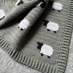 May include: A sage green crochet blanket featuring a repeating pattern of white sheep with black faces and legs. The blanket has a ribbed border and is folded, showcasing the texture and design. The blanket is made of soft yarn.