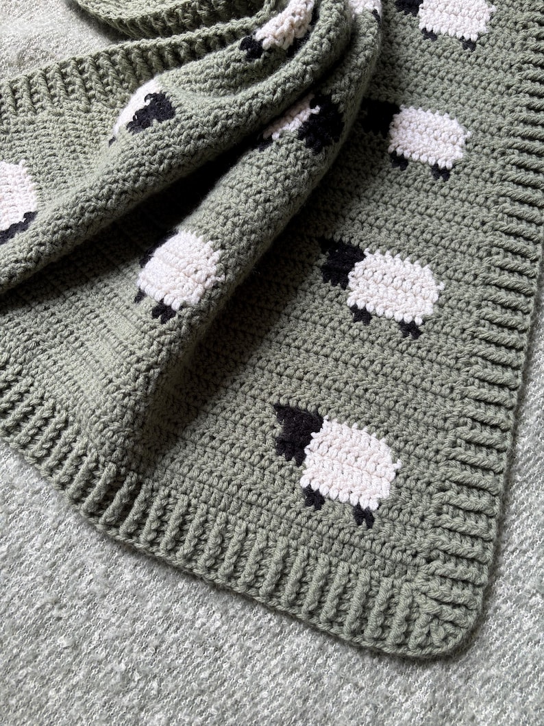May include: A sage green crochet blanket featuring a repeating pattern of white sheep with black faces and legs. The blanket has a ribbed border and is folded, showcasing the texture and design. The blanket is made of soft yarn.