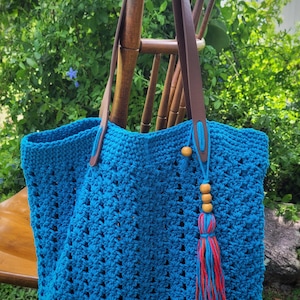 May include: A large, turquoise crochet tote bag with brown leather handles. The bag features a decorative tassel with wooden beads and red and blue threads. The bag is displayed on a wooden chair against a backdrop of green foliage.