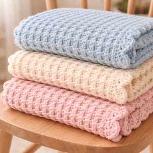 Può includere: Una pila di tre coperte all'uncinetto piegate, nei colori pastello: azzurro, crema e rosa. Le coperte sono disposte ordinatamente su una sedia di legno, mostrando la loro trama e i bordi smerlati. Le coperte appaiono morbide e accoglienti.