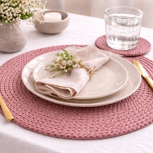 Peut inclure: Mise en place avec un set de table rose tissé, deux assiettes blanches, une serviette en lin pliée et des couverts dorés. Un verre d'eau et un dessous de verre assorti sont visibles. Un petit vase avec des fleurs blanches et une bougie complètent la scène.