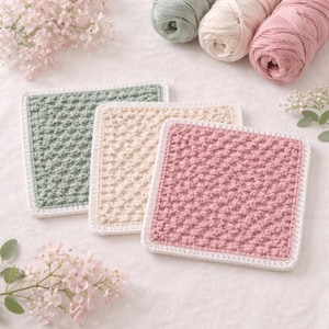 May include: Three square crochet coasters in pastel colors: sage green, cream, and pink. Each coaster has a textured stitch pattern and a white border. Yarn skeins in coordinating colors are in the background, suggesting a handmade craft theme.