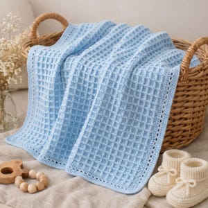 May include: A light blue crocheted blanket with a waffle stitch pattern and a decorative border, accompanied by a pair of cream-coloured knit booties and a wooden bead rattle, all resting inside a woven wicker basket with two handles.