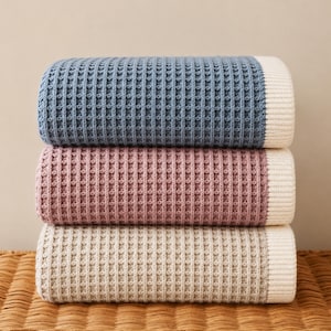 May include: A stack of three folded blankets featuring a textured waffle knit pattern. Each blanket has a solid coloured centre in blue, dusty rose, or beige, and a contrasting cream-coloured ribbed border along the right edge of the fabric.
