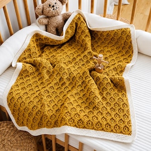 Könnte beinhalten: Eine senfgelbe Häkeldecke mit strukturiertem Wabenmuster und einem einfarbigen cremefarbenen Rand. Die Decke liegt in einem Holzbettchen, zusammen mit einem kleinen braunen Plüschbären und einer winzigen, beweglichen Holzfigur.