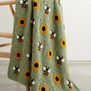 May include: A crochet blanket made of sage green square panels, each featuring either a yellow sunflower with a dark brown centre or a black and yellow bumblebee with white wings. The blanket is constructed from individual squares joined together.