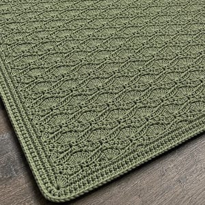 May include: A sage green rectangular crochet blanket featuring a repeating fan-like stitch pattern arranged in diagonal rows. The textile has a dense, textured surface with a uniform border of single crochet stitches framing the intricate design.