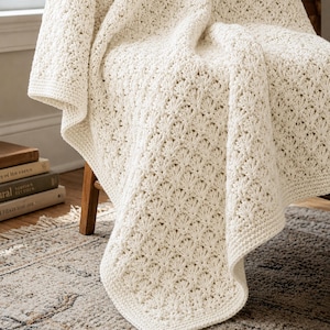 May include: A cream-colored crochet blanket featuring a repeating shell stitch pattern. The textile has a textured surface with a consistent, intricate design throughout, finished with a simple border along the edges of the fabric.