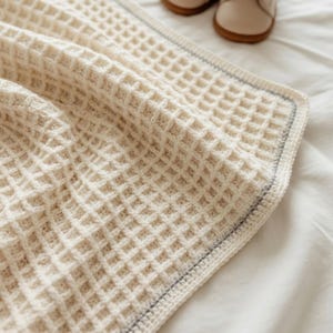 Peut inclure: Une couverture en tricot gaufré de couleur crème présentant un motif à grille texturé. La couverture est finie avec une bordure simple qui inclut un fin détail de ligne grise subtile le long du bord, offrant un aspect propre et structuré.