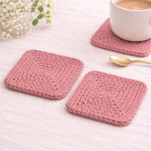 Könnte beinhalten: Drei quadratische, gestrickte Untersetzer in Staubrosa werden gezeigt. Ein Untersetzer hat eine weiße Tasse mit einer braunen Flüssigkeit darauf. Neben der Tasse liegt ein goldener Löffel. Die Untersetzer liegen auf einer weißen Oberfläche.
