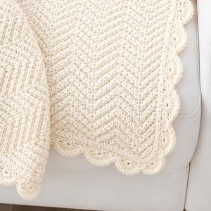 Può includere: Una coperta all'uncinetto color crema caratterizzata da un motivo a chevron testurizzato su tutto il tessuto. I bordi della coperta sono rifiniti con un bordo smerlato decorativo, creando un design arrotondato a conchiglia lungo il perimetro.