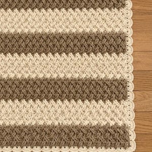 May include: A crocheted blanket featuring alternating horizontal stripes of light brown and cream yarn. The blanket is worked in a textured cable stitch pattern and is finished with a decorative scalloped edge border in a solid cream color.