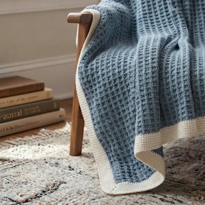 Puede incluir: Una manta de punto gofrado azul con un borde sólido de color crema. La manta está colocada sobre una silla de madera, mostrando un patrón de rejilla texturizado en toda la tela y un borde suave y contrastante a lo largo del perímetro.