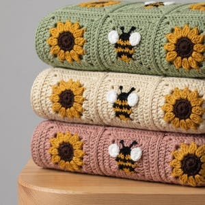 May include: A stack of three crocheted blankets featuring square motifs. Each blanket displays a repeating pattern of yellow sunflowers with brown centres and black and yellow bees with white wings, in sage green, cream, and dusty rose colourways.