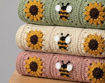 Puede incluir: Una pila de tres mantas de ganchillo con motivos cuadrados. Cada manta muestra un patrón repetitivo de girasoles amarillos con centros marrones y abejas negras y amarillas con alas blancas, en tonos verde salvia, crema y rosa empolvado.