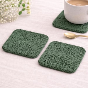 Puede incluir: Un juego de tres posavasos cuadrados de ganchillo verde bosque. Uno de los posavasos tiene una taza blanca con café. Una cuchara dorada está al lado de los posavasos. Los posavasos están sobre una superficie blanca con una ramita de flores blancas.