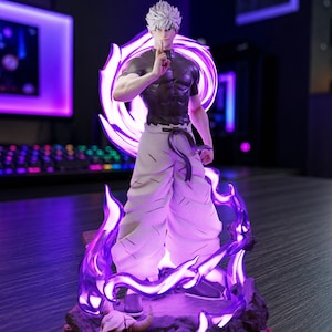 May include: A detailed anime character figurine featuring a figure with spiky white hair, a black t-shirt, and textured white trousers. The figure stands on a base with a skull, surrounded by glowing purple energy effects and Japanese text on the front.