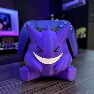 Può includere: Un supporto per controller a forma di Gengar viola, caratterizzato da un ampio sorriso bianco con denti visibili e occhi neri. Il supporto è progettato per sostenere un controller da gioco viola scuro, posizionato dietro le orecchie.