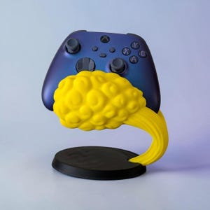 Può includere: Un controller per videogiochi iridescente blu scuro e viola poggiato su un supporto giallo a forma di nuvola. Il supporto presenta una nuvola gialla testurizzata e gorgogliante fissata a una solida base circolare nera per il controller.