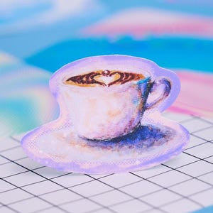 Puede incluir: Una pegatina de estilo acuarela de una taza de café con un diseño en forma de corazón en la espuma. La taza blanca tiene un asa azul y se apoya en un platillo a juego. La pegatina tiene un borde lila claro.