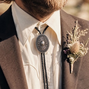 May include: A bolo tie featuring an oval silver-toned metal centerpiece with a sunburst pattern and beaded border, attached to a black braided cord. The tie is worn over a cream-colored button-down shirt under a dark brown blazer with a floral boutonniere.