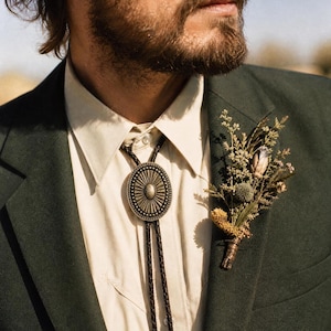 May include: A bolo tie featuring an oval-shaped antique brass-toned metal slide with a sunburst pattern and a central raised oval, attached to a dark brown braided leather cord. A dried floral boutonniere is pinned to the dark green suit jacket.