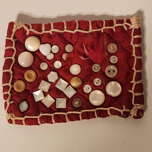 May include: A collection of various buttons displayed on a quilted red fabric. The buttons are in different shapes, sizes, and colors, including white, pearl, and brown. The fabric is stitched with a beige thread.