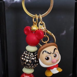 May include: A gold-toned metal keychain featuring a Grumpy character charm with a brown hat, white beard, and red shirt. The keychain includes a red Mickey Mouse-shaped bead, a black glittery spherical bead, and a red marbled bead on a gold ring.