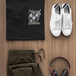 May include: Black t-shirt featuring a record graphic and the text "Pharrell Williams." Accompanied by olive green joggers, white trainers, and maroon headphones. All items are displayed on a wooden surface.