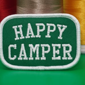 May include: A green patch with the words "HAPPY CAMPER" in white, outlined in white stitching. The patch is rectangular with rounded corners. Spools of red, tan, and yellow thread are in the background.