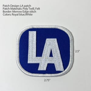 May include: A royal blue and white LA patch made of poly twill and felt with a merrow edge stitch. The patch is rectangular with rounded corners and measures 2.75 inches in width by 2.5 inches in height, featuring the letters LA in white.