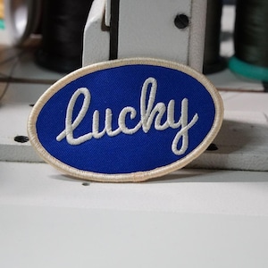May include: An oval-shaped patch with a blue background and the word "Lucky" embroidered in white script. The patch has a cream-colored border. The patch is likely for clothing or accessories.