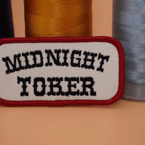 May include: A white embroidered patch with the words "MIDNIGHT TOKER" in black lettering, outlined in red. The patch is rectangular with rounded corners. Spools of blue, gold, and silver thread are in the background.