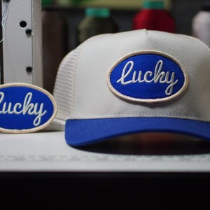 May include: A white and blue trucker hat with a blue brim and a mesh back. The hat features a blue oval patch with the word "Lucky" embroidered in white script. A matching patch is next to the hat.