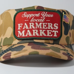 May include: A camouflage baseball cap featuring a rectangular red patch with a dark green border. The patch displays the white embroidered text Support Your Local FARMERS MARKET. A small American flag pin is attached to the side of the cap.