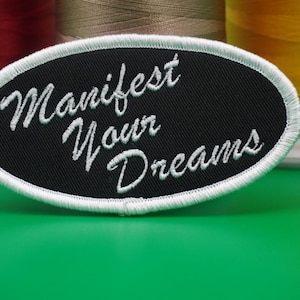 May include: Black oval patch with white embroidered text reading "Manifest Your Dreams." The patch has a white stitched border. Spools of red, tan, and yellow thread are in the background on a green surface.