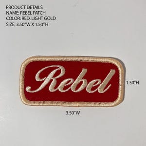 May include: A rectangular embroidered patch with rounded corners featuring a red background and the word Rebel in light gold script. The patch has a light gold border and measures 3.50 inches wide by 1.50 inches high as indicated by text.