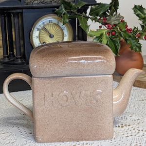 Könnte beinhalten: Beige Keramik-Teekanne mit quadratischem Körper und rundem Deckel. Das Wort "Hovis" ist auf der Vorderseite geprägt. Sie hat einen gebogenen Griff und einen Ausguss. Im Hintergrund eine Uhr und Grün.