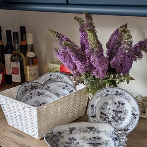 Puede incluir: Una cesta de mimbre blanca contiene varios platos con diseños en blanco y negro. Una fuente a juego y platos adicionales están colocados sobre una superficie de madera. Un jarrón con flores moradas y botellas de vino están en el fondo.