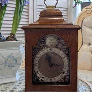 May include: A brown wooden mantel clock with a brass handle and a round clock face featuring Roman numerals. The clock has decorative detailing and sits on a table. The clock has a small inscription at the top.