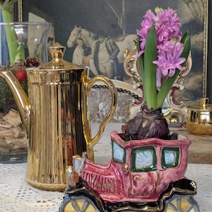 May include: A gold-coloured, ribbed teapot and a pink ceramic car-shaped planter holding purple hyacinth flowers. The car has black accents and yellow-trimmed wheels. A small gold sugar bowl is also visible.