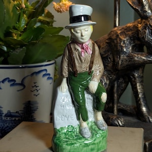 May include: A ceramic figurine of a young boy in a top hat, brown jacket, green pants, and suspenders, seated on a stone. The stone reads "30% Miles to Dove." The figurine has a pink scarf and gray shoes. The base is green with grass.