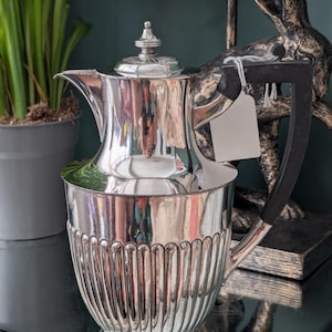 May include: A silver-colored pitcher with a black handle and a lid. The pitcher has a fluted design on the lower portion and a spout for pouring. A blank tag hangs from the handle. The pitcher is likely for serving beverages.