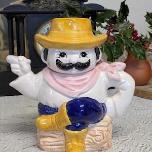 Może przedstawiać: Ceramiczny czajnik w stylu kowbojskim. Czajnik jest biały z żółtym kapeluszem, czarnym wąsem i niebieskimi spodniami. Ma różową chustę i żółte buty. Czajnik stoi na drewnianej podstawie.