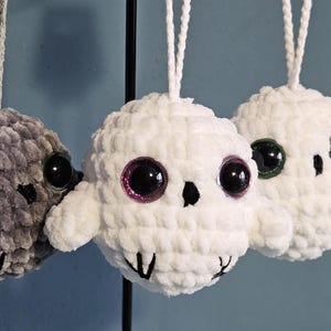 Pode incluir: Três enfeites de caveira de crochê feitos à mão em cinza e branco, cada um com olhos grandes e brilhantes. A caveira cinza tem olhos verdes, enquanto as caveiras brancas têm olhos rosa e verdes. Cada enfeite tem uma alça para pendurar.