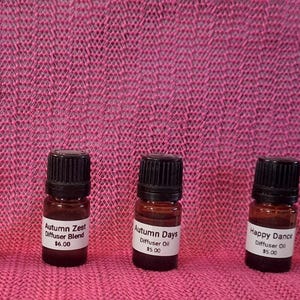 May include: A row of small amber glass bottles with black caps, each labeled with a different scent name. The labels include "Mulled Cider," "Winter Sunshine," "Autumn Zest," "Autumn Day," "Happy Dance," "Unwind," and "Morning Blossom." The bottles are arranged on a pink textured surface.