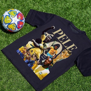 May include: A navy blue t-shirt featuring a collage of images of Pel&eacute;, a famous soccer player, with the word "PELE" in gold lettering. A soccer ball with colorful panels is in the background.