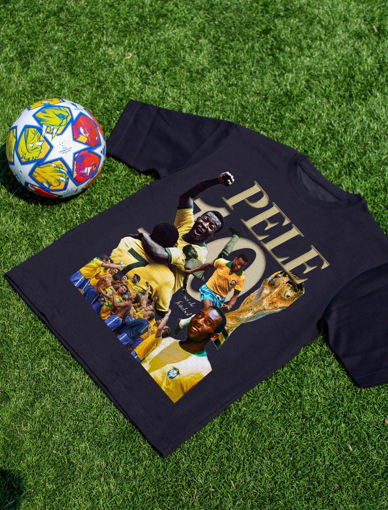 May include: A navy blue t-shirt featuring a collage of images of Pel&eacute;, a famous soccer player, with the word "PELE" in gold lettering. A soccer ball with colorful panels is in the background.