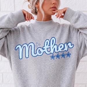 May include: A heather gray long-sleeve sweatshirt with a mock neck and ribbed cuffs. The front features the word Mother in a blue script font with a distressed texture, accompanied by several small blue star outlines and four solid blue stars.